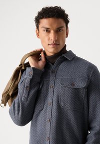 Gray herringbone patterned shirt with brown buttons, two chest pockets, and a collar, paired with a light brown bag.