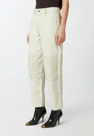 Pantalones color crema con acentos de tachuelas en la parte inferior de las piernas, con un corte recto y bolsillos frontales, combinados con botas negras de tobillo puntiagudas.