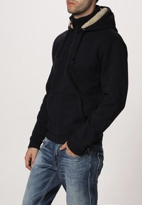 Uomo che indossa un hoodie nero con zip e cappuccio foderato in sherpa, con jeans blu sbiaditi, in piedi con una mano in tasca.