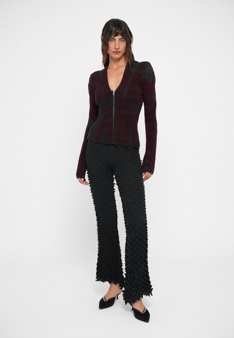 Pull zippé bordeaux foncé avec un motif carré, associé à un pantalon noir évasé texturé présentant des bords scallopés proéminents.