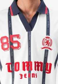Close-up van een wit polo shirt met marinekleurige kraag, een rode "85" patch, rode tekst "Tommy Jeans", verticale marineblauwe strepen met "tommy jeans" en een rode leeuwenkroon.