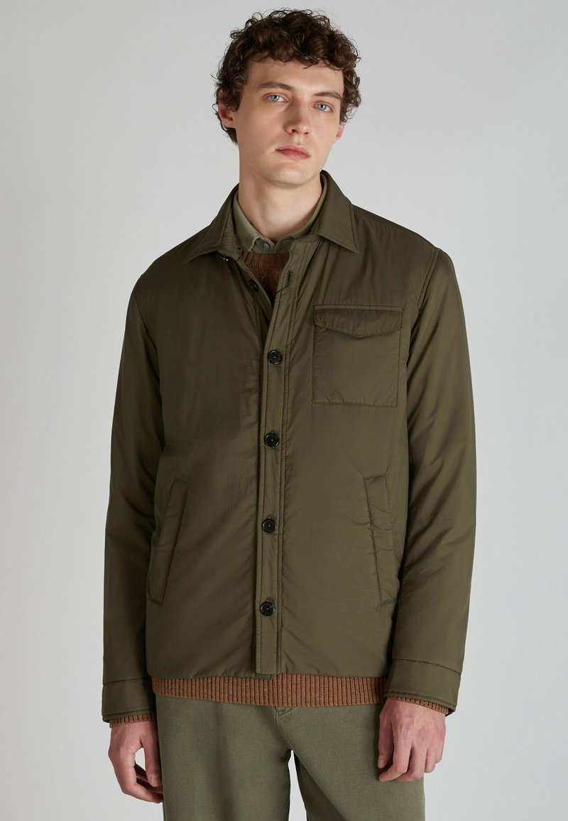 Chaqueta abotonada verde oliva con cuello de camisa, bolsillos frontales y botones negros. Textura suave, diseño acolchado y corte relajado.