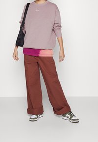 Světle fialová mikina s bílým logem Nike, navrstvená přes tričko s růžovým gradientem. Hnědé široké kalhoty a zeleno-bílé tenisky dotvářejí celý outfit.