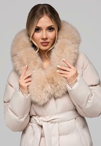 Bezešvý béžový kabát s velkým límcem z umělé kožešiny. Obsahuje pásek v pase a hladkou texturu. Ruce umístěné na límci.