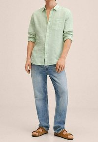 Chemise en lin vert clair avec des boutons sur le devant et une poche poitrine, associée à un jean en denim bleu clair et des sandales ouvertes marron.