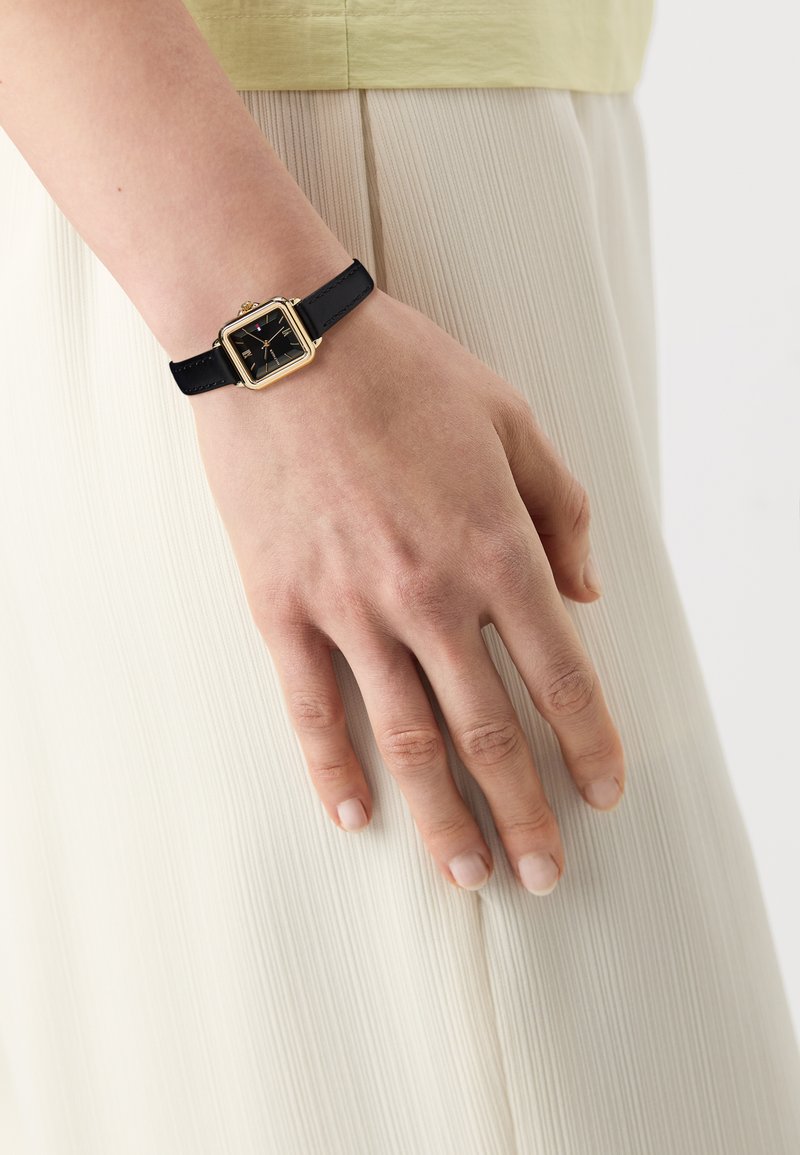 Montre à cadran carré avec un bracelet en cuir noir, lunette dorée et cadran noir affichant des index et aiguilles dorés, portée sur une main.