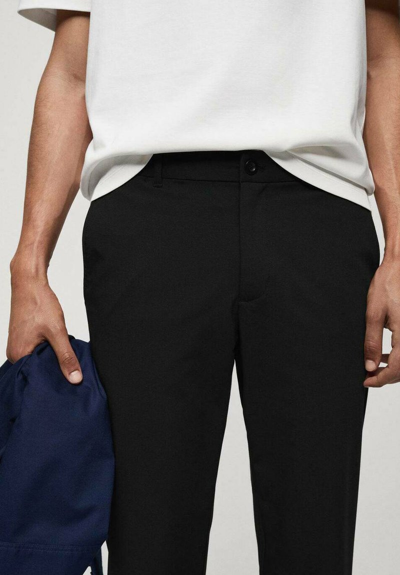 Pantalon noir en tissu lisse, offrant une coupe plate à l'avant, des poches latérales et une fermeture à bouton à la taille. Porté avec une chemise blanche.