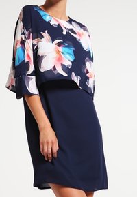 Šaty v námořnické barvě s květinovým potiskem a volnými krátkými rukávy. Vrchní část má živý vzor květin v růžové, modré a bílé barvě na hladké látce.