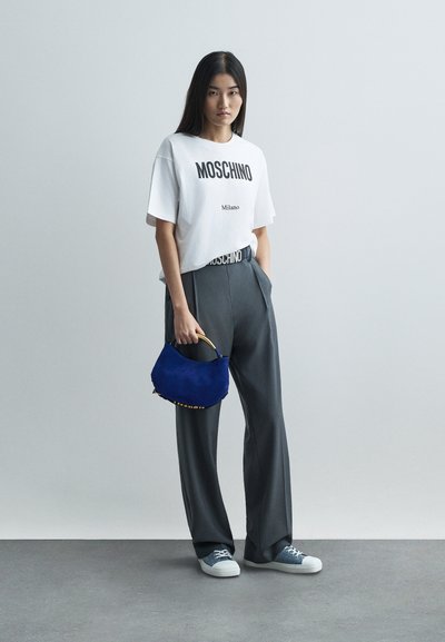 Mladá žena v bílé tričku Moschino, šedých širokých kalhotách, modrých teniskách, držící malou modrou kabelku na jednolitém šedém pozadí.