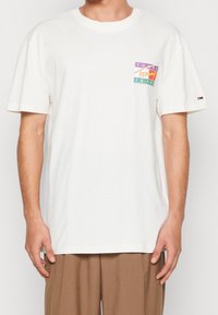 Camiseta de algodón blanca de manga corta, con un parche de logo colorido en púrpura, verde y naranja en el lado izquierdo del pecho.