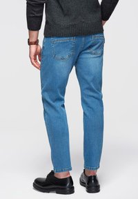 Jean en denim bleu clair avec une coupe droite, doté de poches arrière et d'un effet délavé, associé à des chaussures noires et un pull sombre.