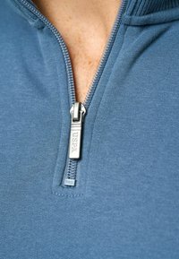 Kék cipzáras pulóver puha anyagból, ezüst színű cipzárral, rajta "USPA" márkajelzéssel és bordázott nyakkivágással. Közelről készült kép.