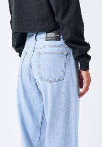 Light blue denim jeans featuring a high-waisted design, back patch pockets, and a black brand label near the waistband.