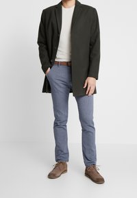 Cappotto lungo verde scuro, maglione beige chiaro e pantaloni blu texturizzati. Scarpe marroni completano l'outfit con una cintura marrone in risalto.