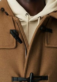 Cappotto di lana marrone con chiusure a toggles nere, dotato di zip e con un hoodie beige indossato sotto. Visto ravvicinato delle texture.