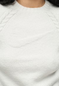 Κοντινό πλάνο ατόμου που φοράει λευκό πλεκτό πουλόβερ με διακριτικό ανάγλυφο λογότυπο Nike Swoosh στο στήθος.