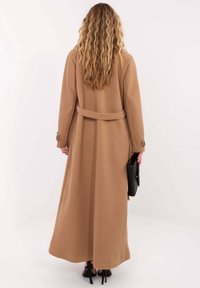 Długi płaszcz w kolorze wielbłądzim z paskiem w talii, charakteryzujący się dopasowanym krojem, zapinanymi na guziki mankietami i gładką fakturą. Widoczna czarna torebka i buty na obcasie.