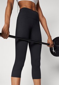 Schwarze, enganliegende Sportleggings mit hoher Taille, cropped Länge und glatter Textur, kombiniert mit einer schwarzen Hantel, die in einer Hand gehalten wird.