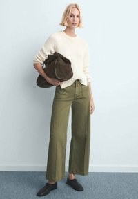 Maglione lavorato a maglia color crema abbinato a pantaloni ampi verde oliva; la modella tiene una borsa morbida marrone scuro; ambientazione minimalista con parete azzurro chiaro.