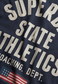 Camiseta de tejido navy con un atrevido texto blanco que dice "SUPERDRY STATE ATHLETICS COACHING DEP." y un pequeño gráfico de la bandera de EE. UU. Diseño suave y casual.