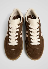 Brune suede sneakers med creme accenter, der har et struktureret design, hvide snørebånd og to vandrette striber i en lysere nuance.