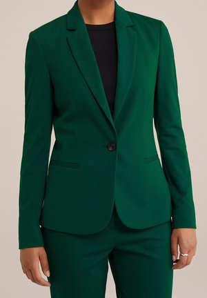 Mujer que lleva un blazer ajustado verde oscuro y pantalones a juego sobre una camiseta negra, con un anillo de plata en la mano derecha.