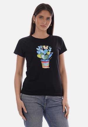 Donna che indossa una t-shirt nera con un design di un cactus a forma di cuore multicolore in un vaso a righe, abbinata a jeans blu, in piedi su sfondo bianco.