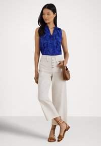 Donna che indossa una blusa senza maniche blu con motivo, pantaloni bianchi a vita alta con bottoni, sandali marroni e tiene una borsa marrone.