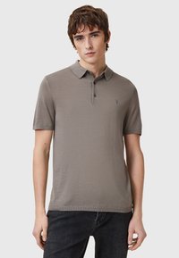 Kortärmad polo i ljus taupe med texturerat tyg, tvåknappsstängning och en mörk broderad logga på bröstet.