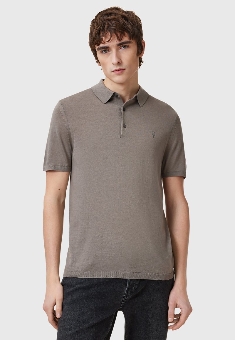 Kortärmad polo i ljus taupe med texturerat tyg, tvåknappsstängning och en mörk broderad logga på bröstet.