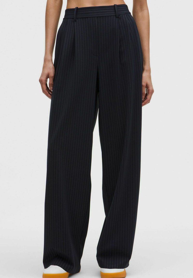 Črne široke hlače z navpičnimi črtami in zankami za pas, nošene z belimi supergami, prikazane od pasu do čevljev na enobarvni podlagi.