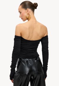 Zwarte off-shoulder top met geplooide lange mouwen; gecombineerd met glanzende zwarte nep krokodillenleren broek met zakken.