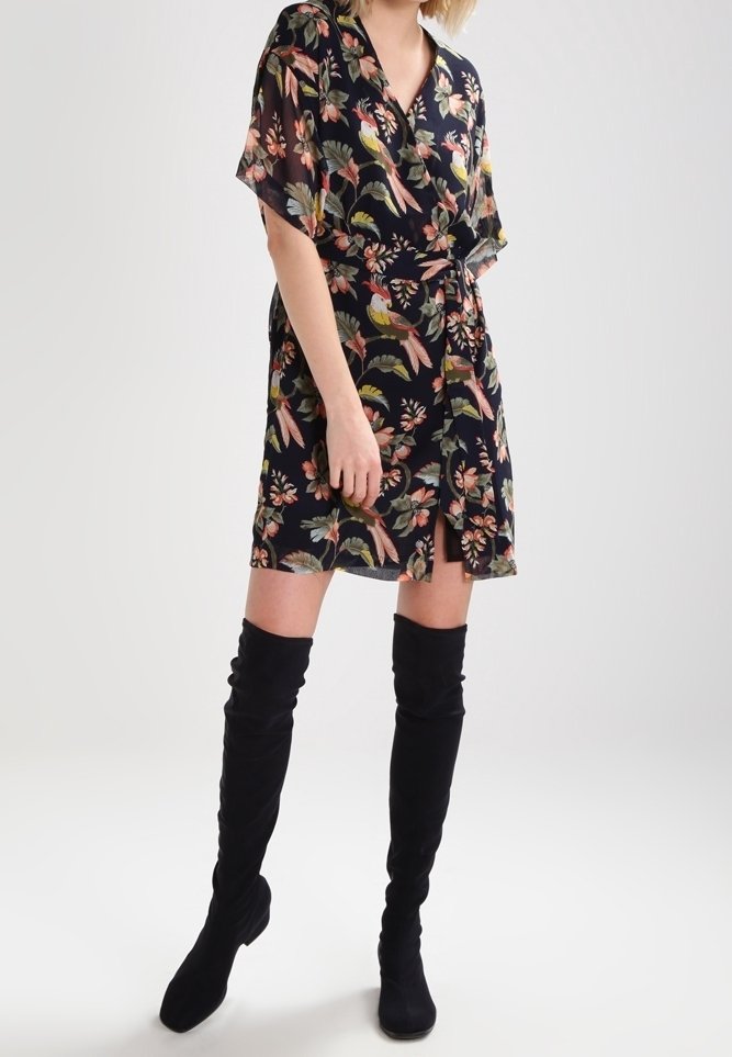 Kvinna i en svart blommig omslagsklänning med färgglada fågel- och blommönster, kombinerad med svarta överknästövlar.