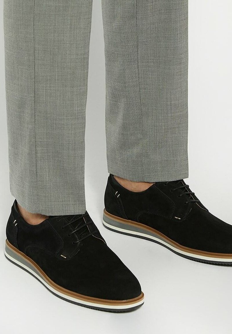 Zwarte suède veterschoenen met een ronde neus en een lichtbruine rubberen zool. Broek in een lichtgrijze geruite patroon.