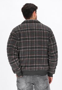 Grau kariertes Flanelljackett mit roten Akzenten, dicke Textur, gerippte Bündchen und einen Kragen. Trägt graue Jeans.