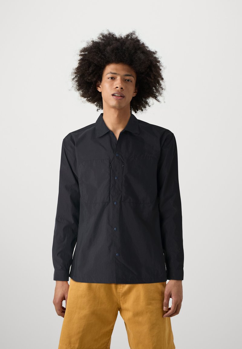 Black long-sleeve shirt with a collar, featuring two chest pockets and blue buttons. Smooth texture, relaxed fit, paired with yellow shorts.