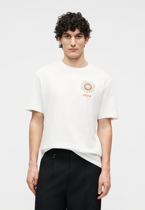 Młody mężczyzna o kręconych ciemnych włosach, ubrany w białą koszulkę z nadrukiem słońca i logo "just cavalli" na piersi, w połączeniu z czarnymi spodniami, na neutralnym tle.