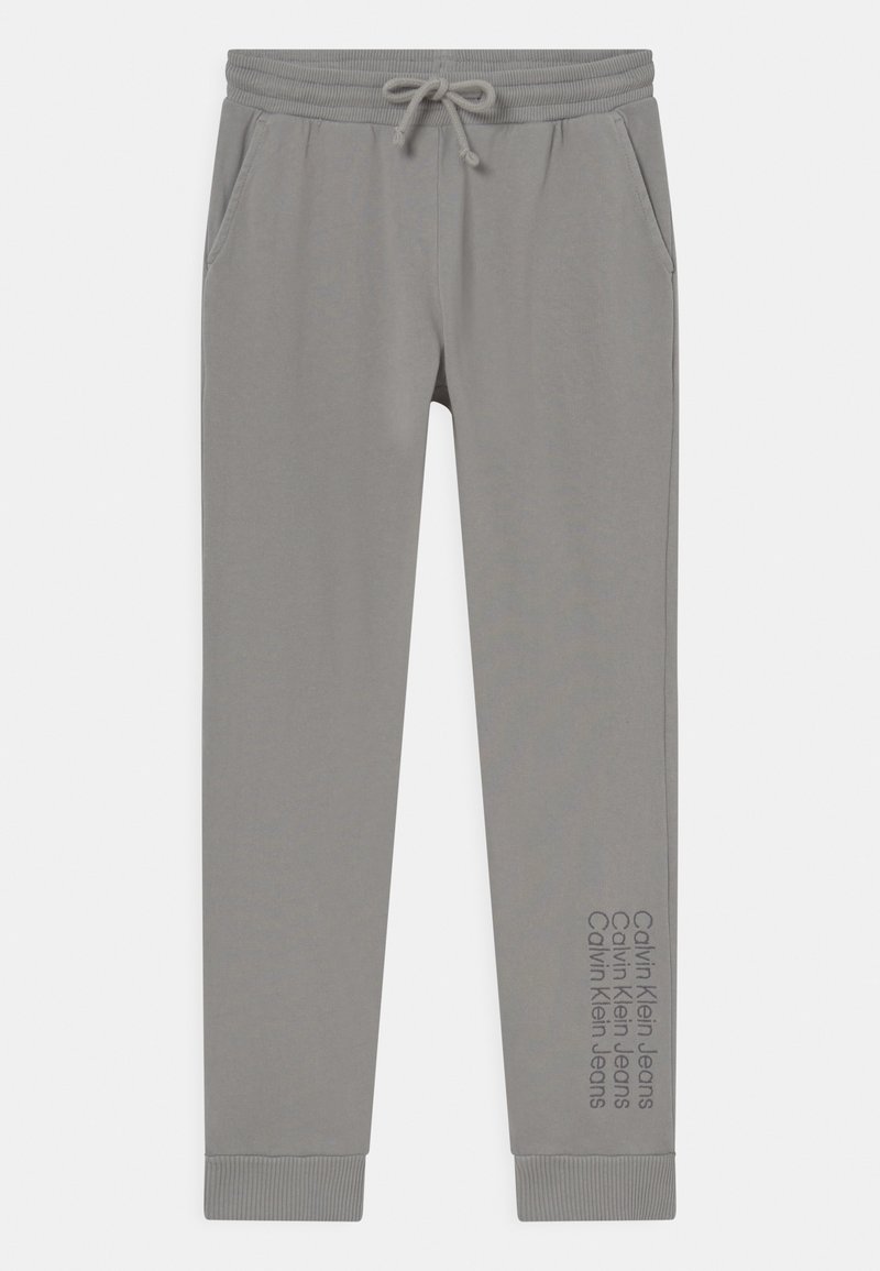 Světlé šedé tepláky s elastickým pasem a nastavovací šňůrkou. Obsahují boční kapsy a nápis "Calvin Klein Jeans" vytištěný na noze.
