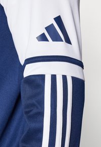 Mornarsko plava i bijela atletska jakna sa rukavima koja ima tri bijele pruge i plavi Adidas logo. Glatka tekstura s sportskim dizajnom.