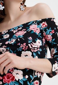 Top bez ramínek vyrobený z černé látky s živými květinovými vzory v růžové, bílé a tyrkysové barvě. Tříčtvrteční rukávy.