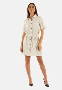 Abito camicia in lino beige con maniche corte, chiusura a bottoni, tasca anteriore e cintura legata in vita, caratterizzato da una vestibilità ampia e orlo sopra il ginocchio.