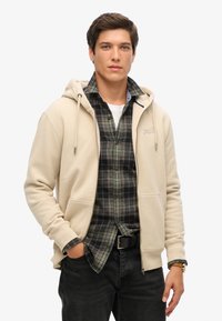 Sudadera beige con capucha y cordones sobre una camisa de cuadro oscurecido. Presenta bolsillos frontales y una textura suave y lisa. Jeans negros y cinturón.