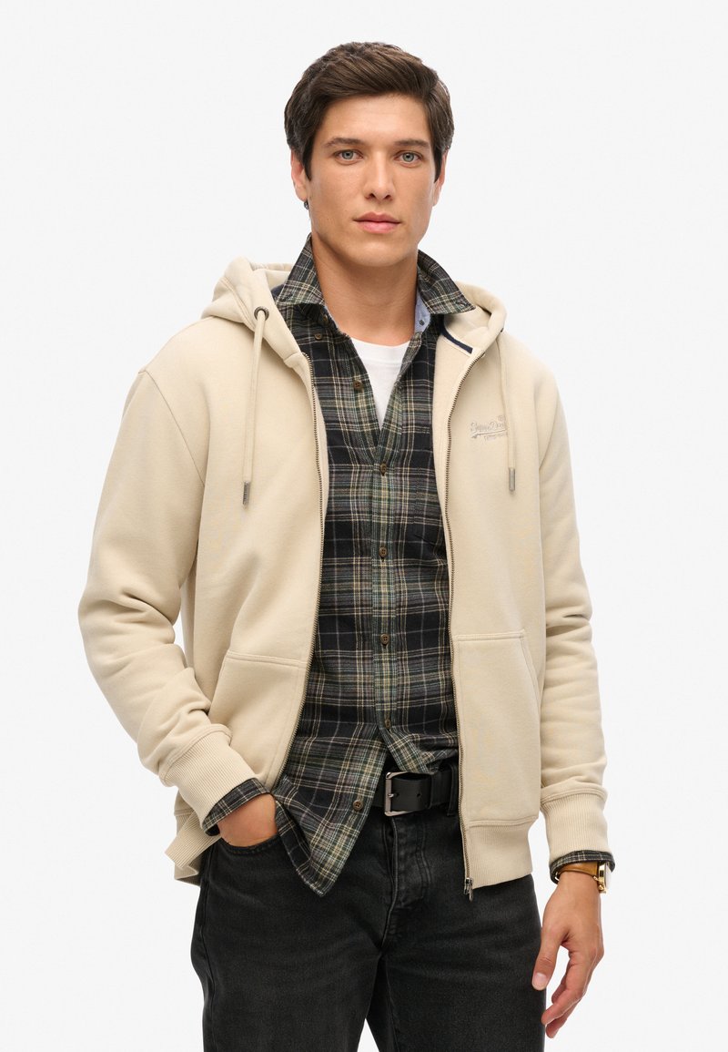 Sudadera beige con capucha y cordones sobre una camisa de cuadro oscurecido. Presenta bolsillos frontales y una textura suave y lisa. Jeans negros y cinturón.
