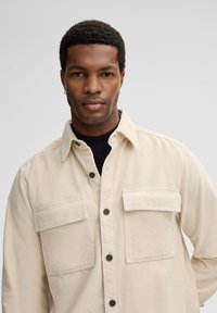 Beige corduroy shirt with a button-up front, two chest pockets, and a standard collar. Textured fabric adds dimension.