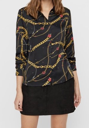 Vrouw draagt een zwarte blouse met lange mouwen, gouden ketting en rode bloemmotieven, gecombineerd met een effen zwarte rok.