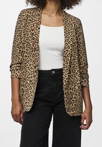 Žena v leopardo vzorovaném blejzru přes bílé tričko a černé kalhoty s vysokým pasem, stojící na jednobarevném světlém pozadí.