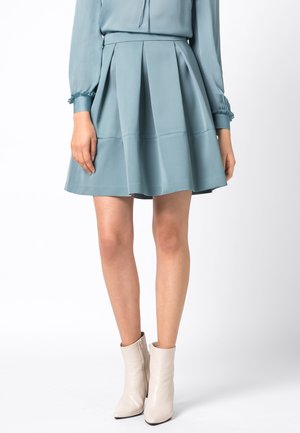 Femme portant une jupe plissée bleu clair, un chemisier à manches longues et des bottines blanches, debout devant un fond blanc.