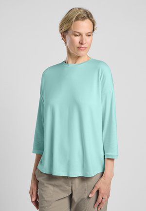 Lichtblauw losvallend topje met driekwart mouwen en een ronde halslijn. Zachte textuur, minimaal ontwerp, gecombineerd met beige broek.