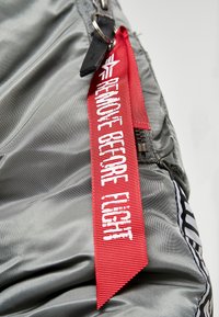 Grå nylonväv med en blank yta har en röd etikett som säger "TA BORT FÖRE FLYGNING." Inkluderar metallbeslag och en dragkedjedetalj.