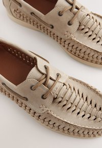 Chaussures en daim beige présentant des détails tissés sur les côtés, des lacets ronds et des œillets noirs. Semelle plate à texture lisse.